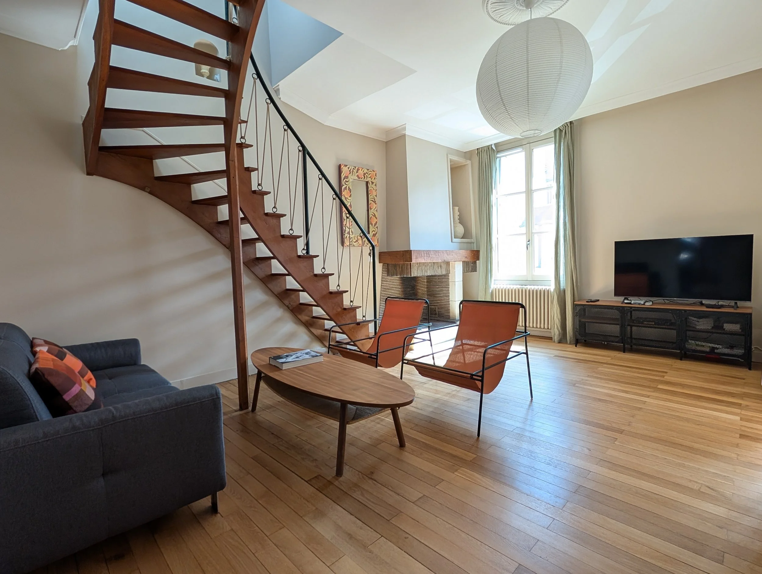 Living room, sofa, wooden table, orange armchairs, fireplace, large window, wooden staircase, hardwood floors