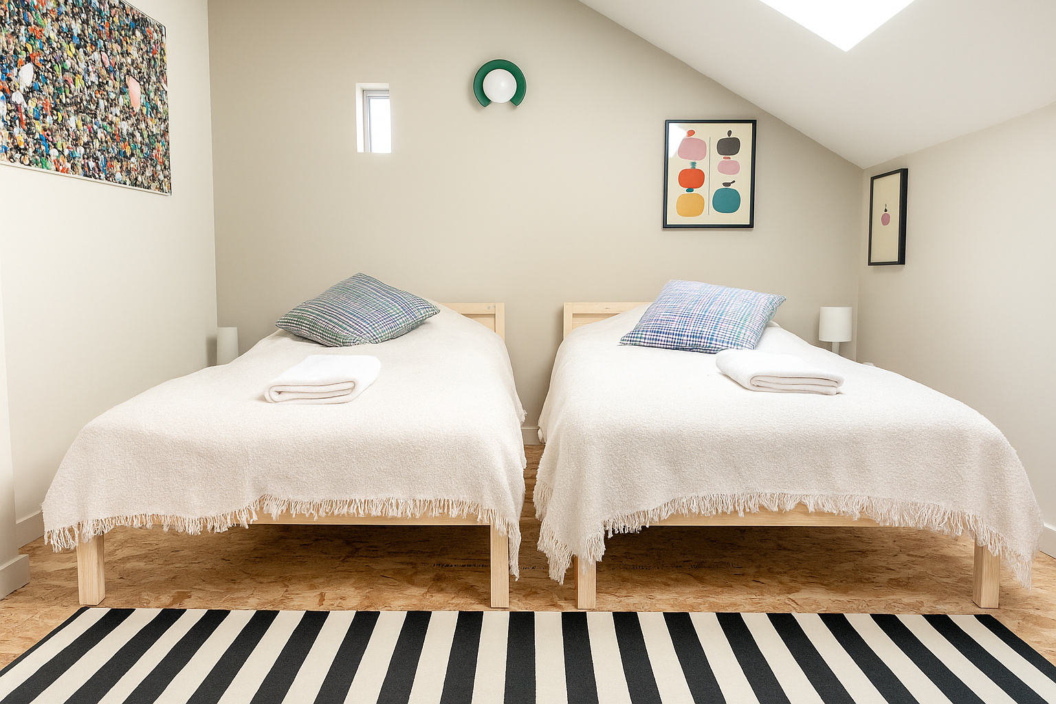 A bedroom with two light-wood single beds, white blankets, checkered pillows, a black-and-white striped rug, colorful artwork, and small windows.