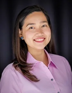 A young woman with long dark hair smiling, wearing a pink button-up shirt and blue earrings against a dark background.