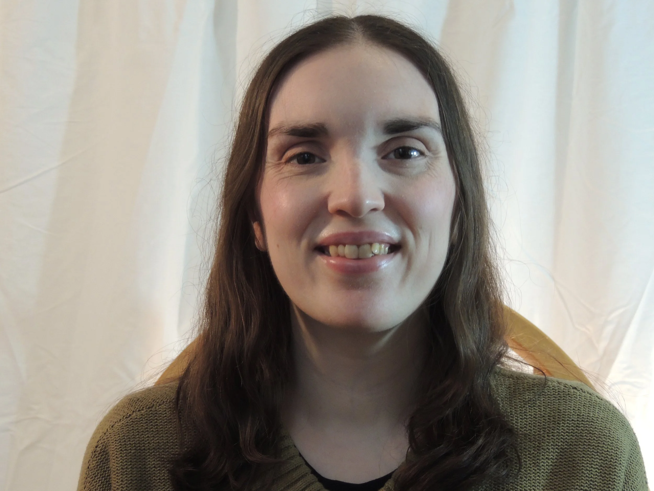A woman with long brown hair, smiling, and wearing a brown sweater, standing in front of light-colored curtains.