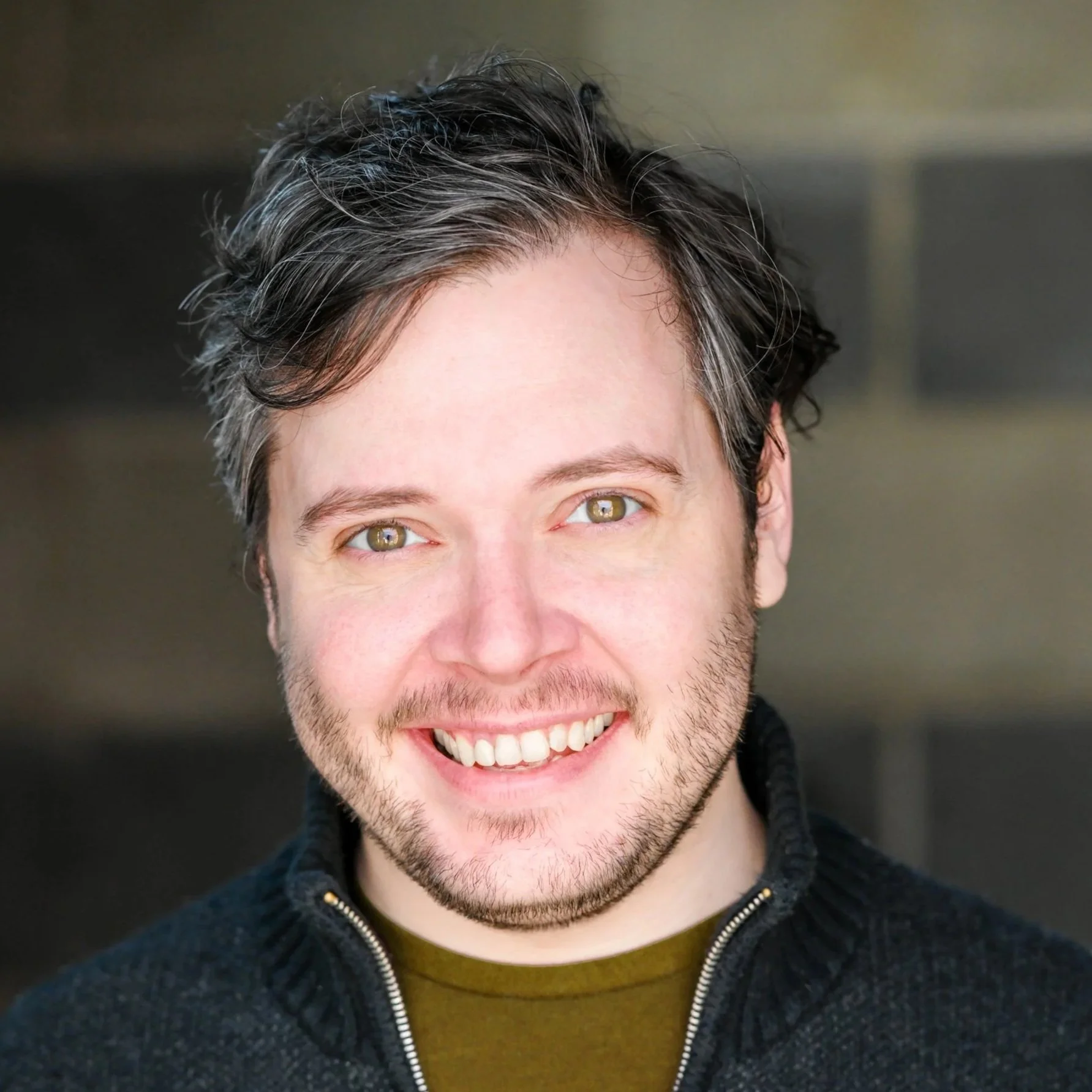 Close-up of a smiling man with short dark hair, green eyes, and a light beard, wearing a black zippered sweater and standing against a blurred background.