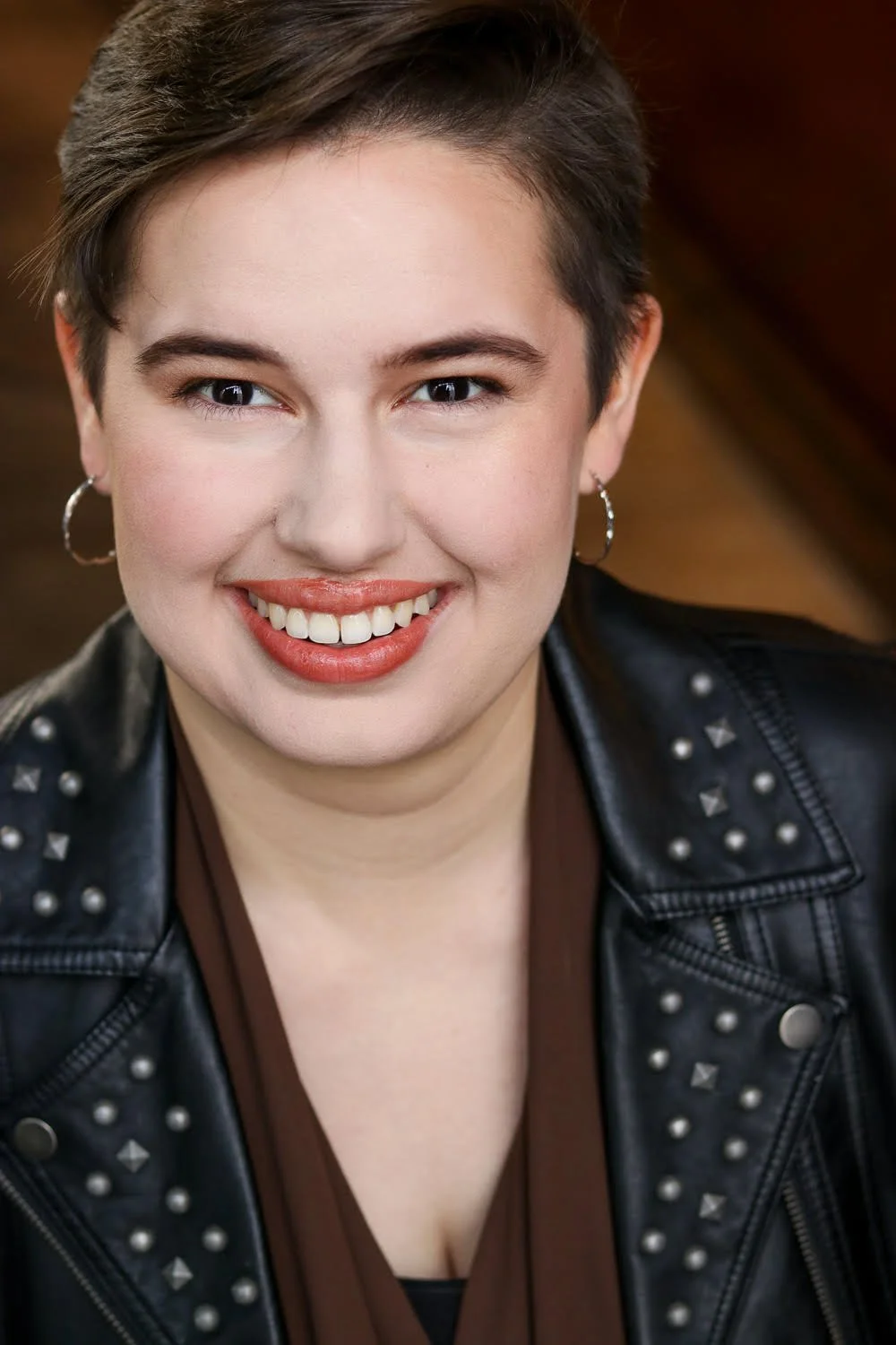 Smiling woman with short dark hair, wearing earrings, a studded black leather jacket, and a brown top.