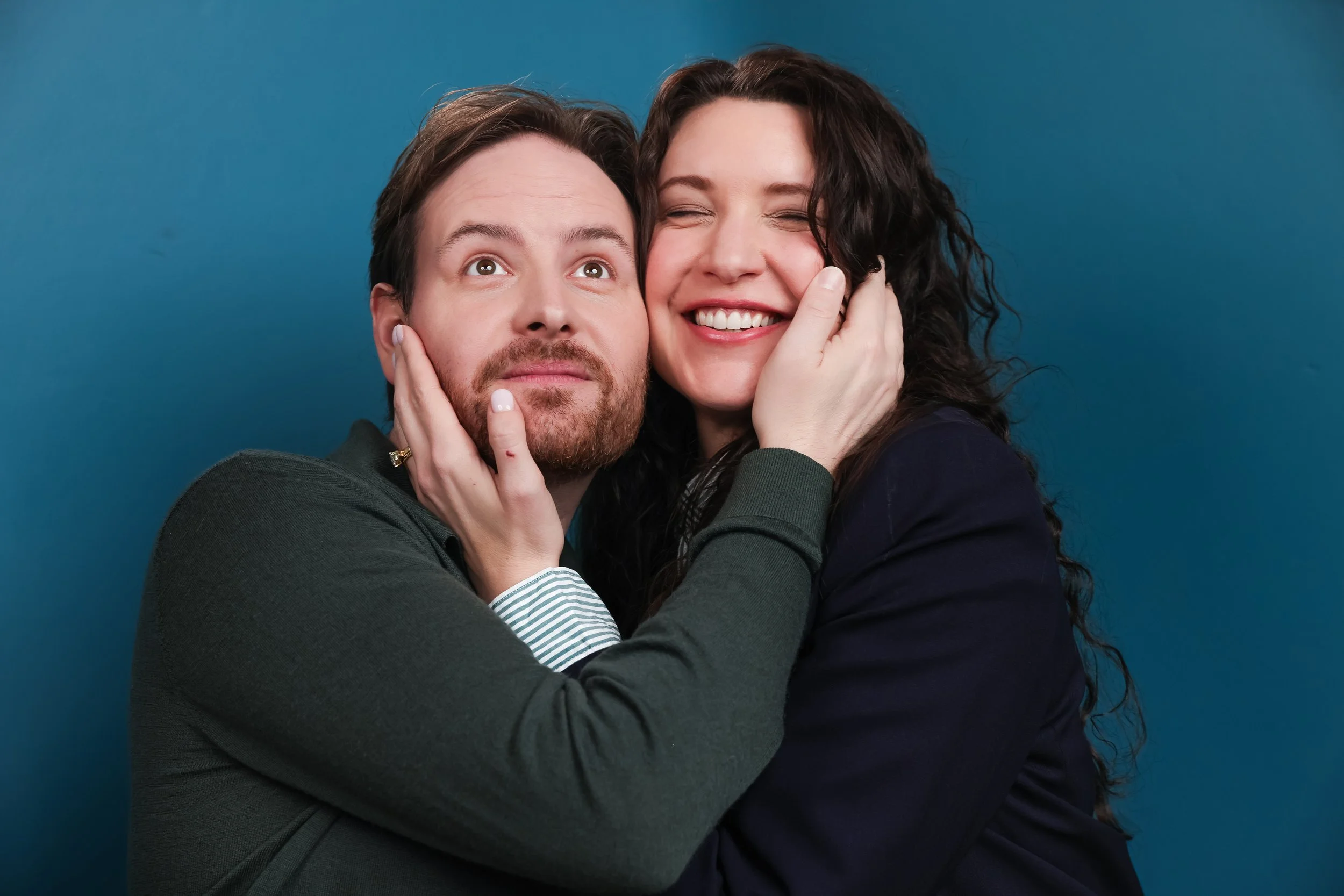 A man and woman hugging and smiling against a blue background, with the woman gently pressing her hand against the man's cheek.