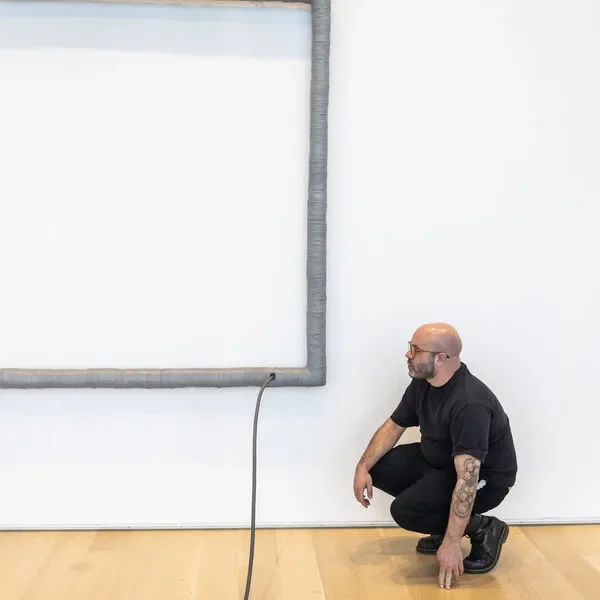 A bald man with glasses and tattoos crouching near a white wall with a large empty whiteboard or screen framed in gray. He is wearing a black t-shirt and black pants.