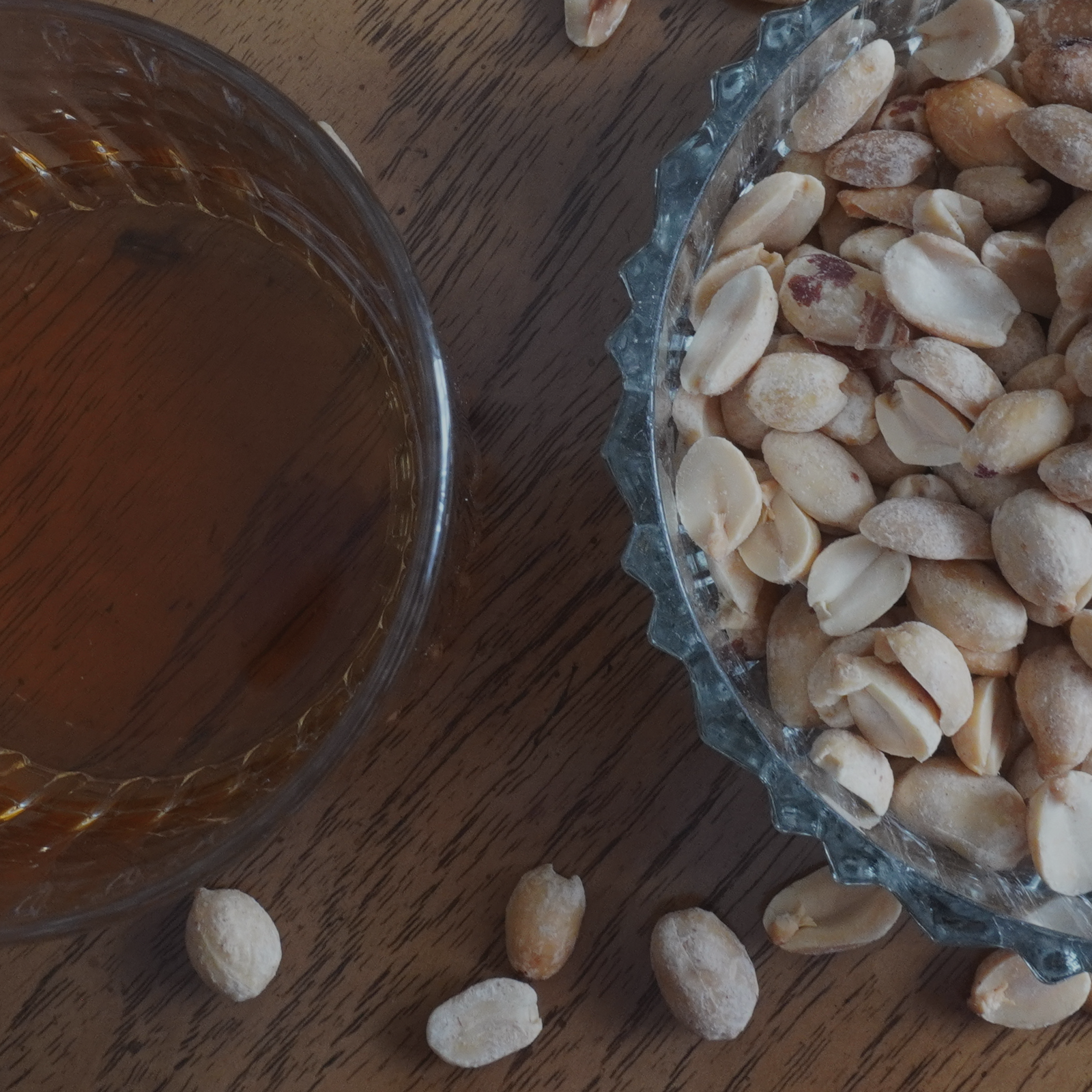 A glass of whiskey next to a dish of peanuts on a wooden table.