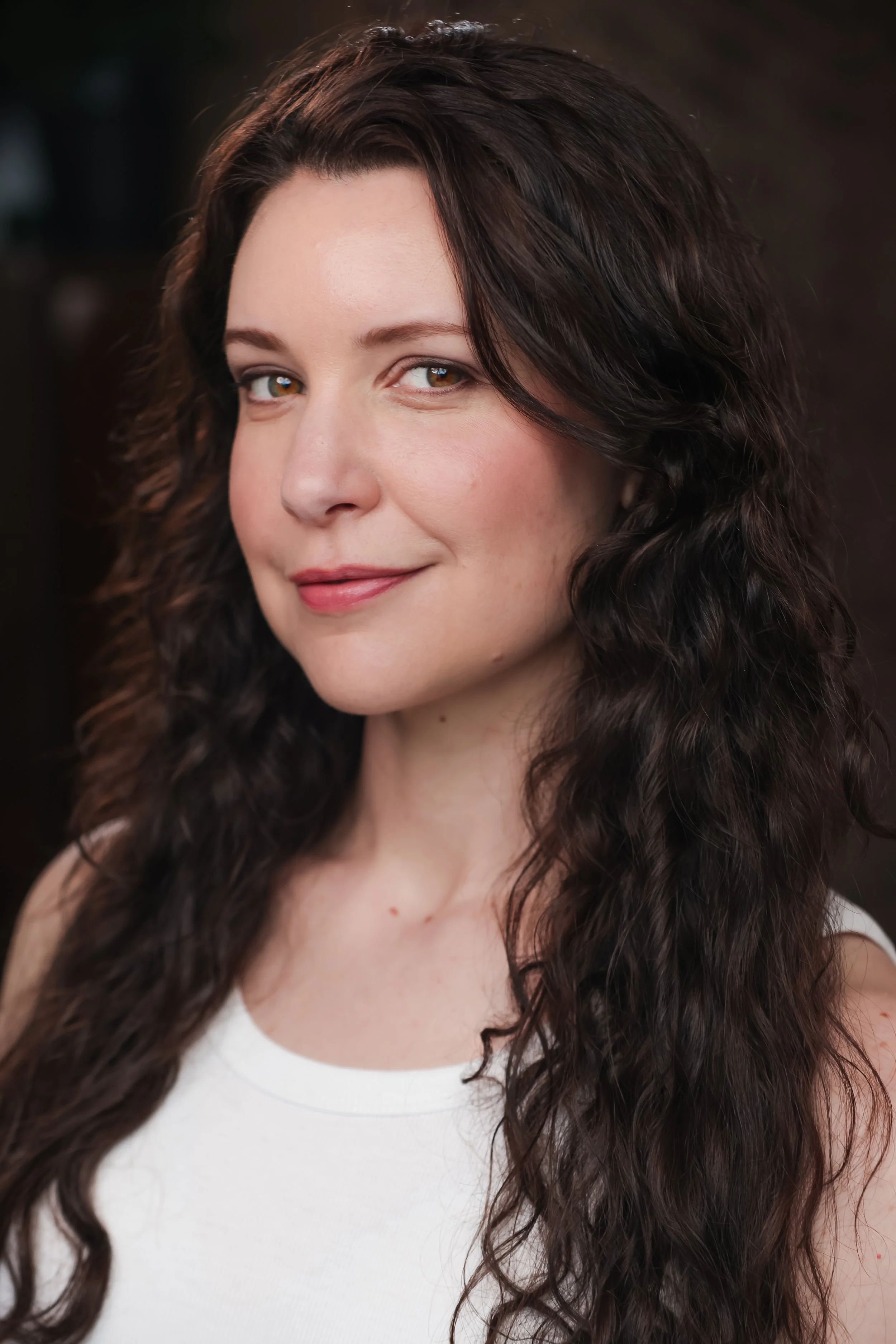 Close-up of a woman with long, wavy dark hair and light skin, wearing a white top, looking at the camera with a slight smile.
