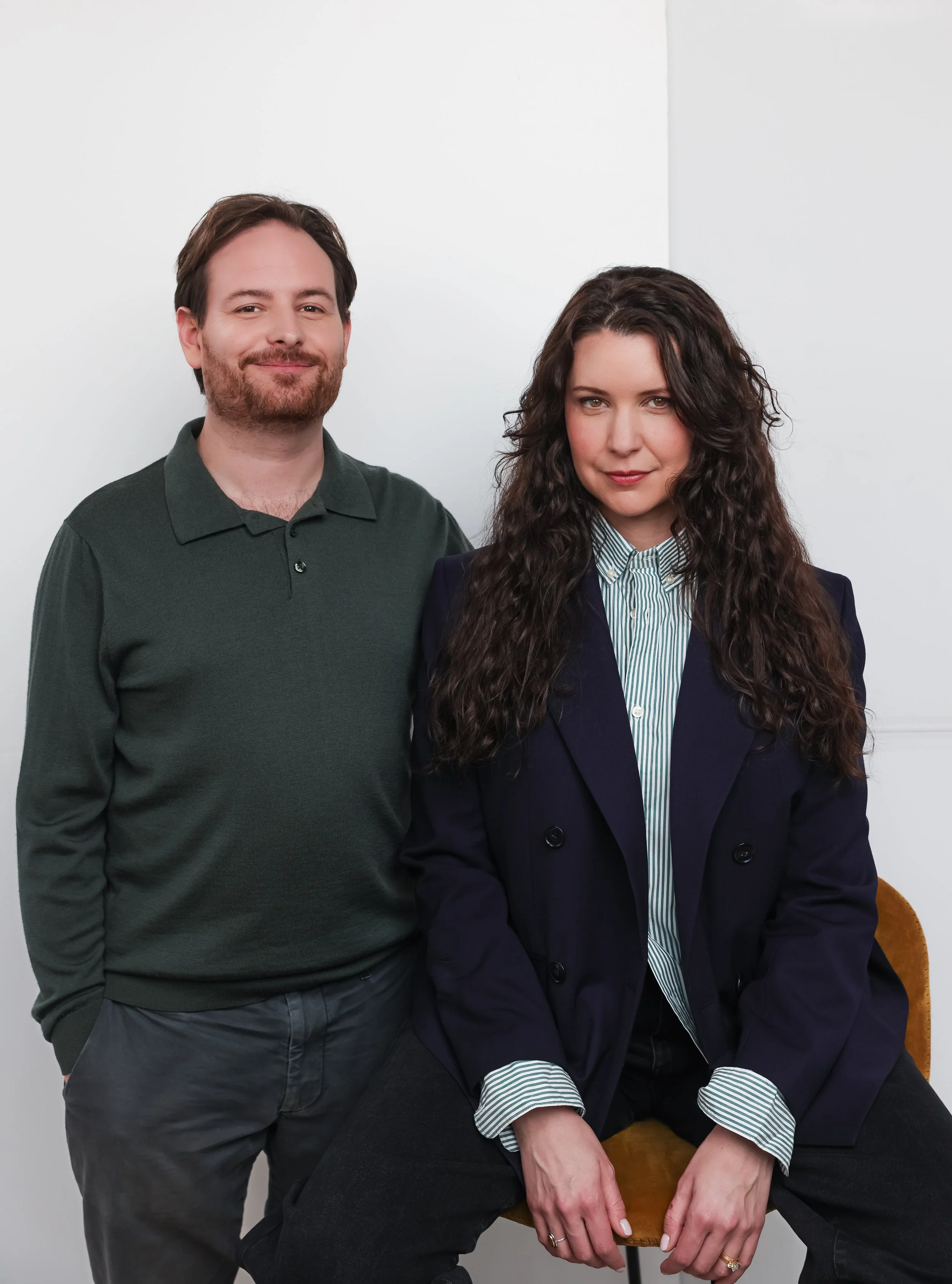 A man and a woman posing together in a room with light-colored walls. The man is standing on the left, wearing a dark green polo shirt, and has brown hair and a beard. The woman is seated on a wooden chair, wearing a dark blazer and a striped shirt, with long, curly dark hair. They are both smiling, and the woman has her hands resting on her lap.