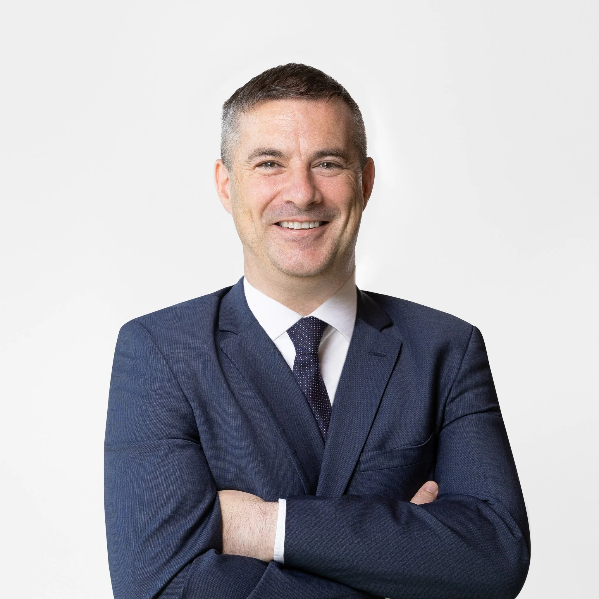 Business man wearing a dark blue suit and white shirt, smiling, with his arms crossed against a plain white background.