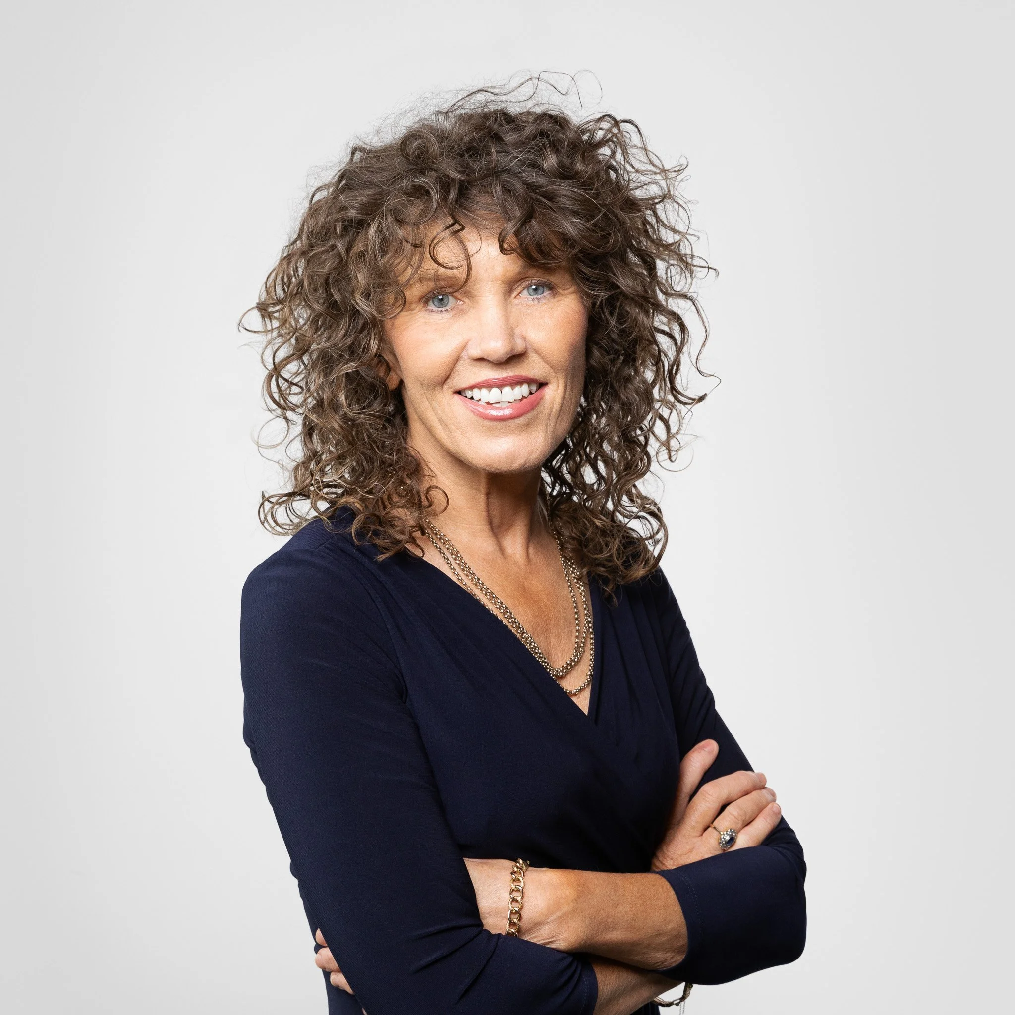 Close-up portrait of Patricia Walley Consultant Solicitor at David Walley & Co Solicitors wearing a dark long-sleeve shirt, layered necklaces, and a ring, with her arms crossed against a plain light gray background.