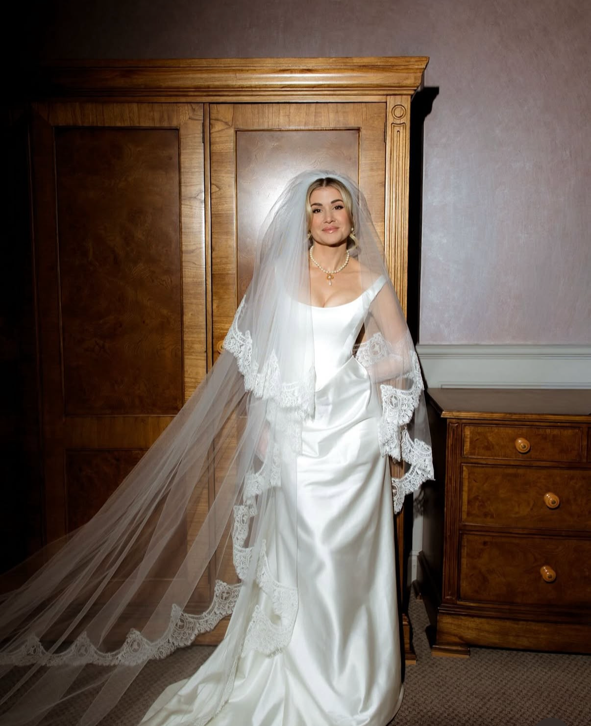 A bride in a satin wedding dress with lace trim, wearing a pearl necklace and veil, standing in front of wooden furniture.