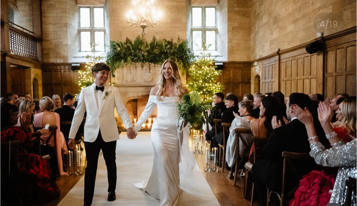 Bride and groom walking hand in hand at their wedding ceremony in a decorated hall with seated guests clapping, Christmas trees with lights, and large windows.