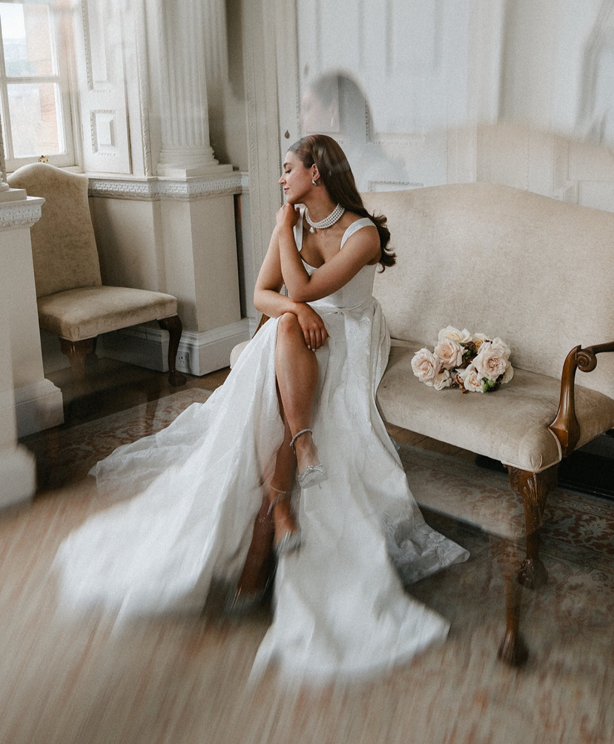 A bride in a white wedding gown sitting on a vintage beige sofa, with her eyes closed and hand on her neck, in a room with classic decor and a bouquet of roses on the sofa.