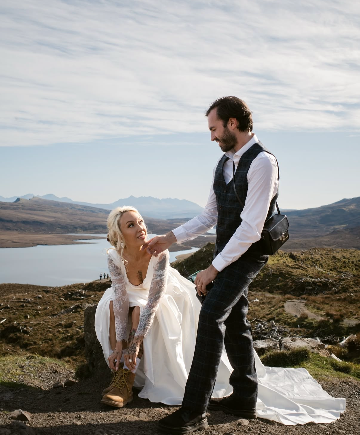 A man and woman outdoors in a scenic mountainous area, with a lake in the background. The woman is sitting on a rock wearing a white dress with lace sleeves, and the man is standing in front of her in a vest and checkered pants, holding her hand and 
