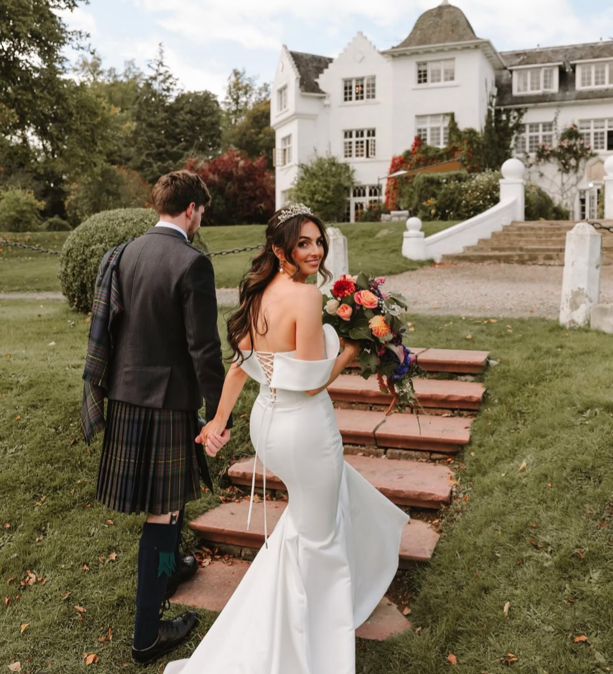 A bride in a white wedding gown holding a colorful bouquet, smiling and turning back, holding hands with a groom in a traditional Scottish kilt and jacket, standing on steps outside a large white house with green trees.