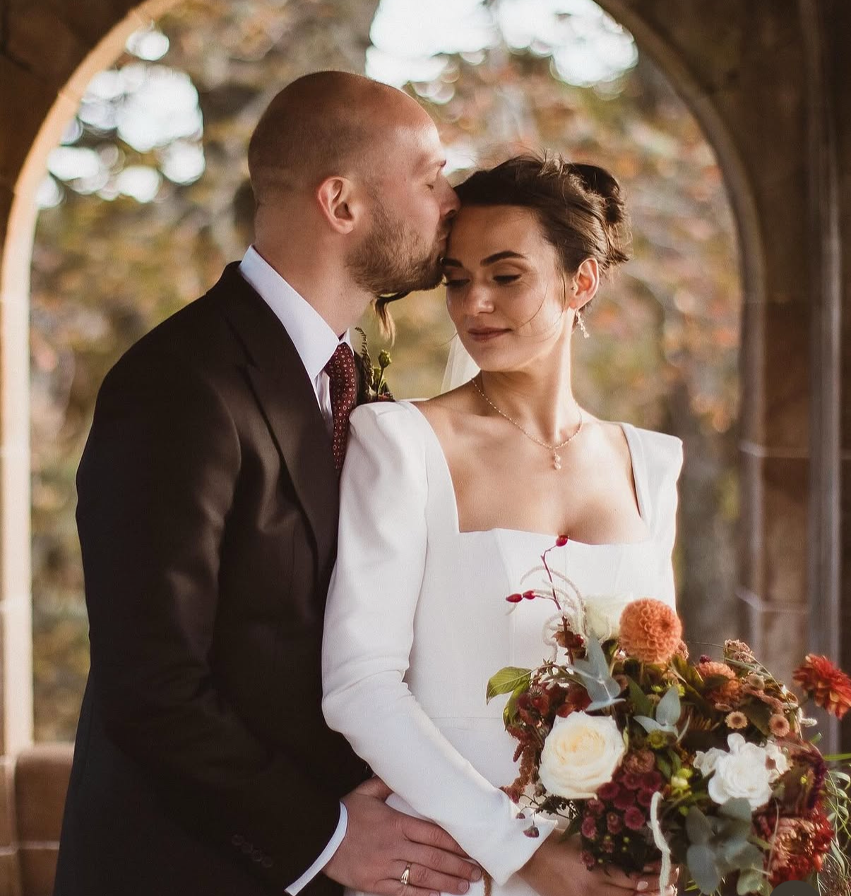 A bride and groom sharing a kiss on their wedding day, with the groom in a black suit and the bride holding a bouquet of flowers, standing under an archway with blurred background of trees.