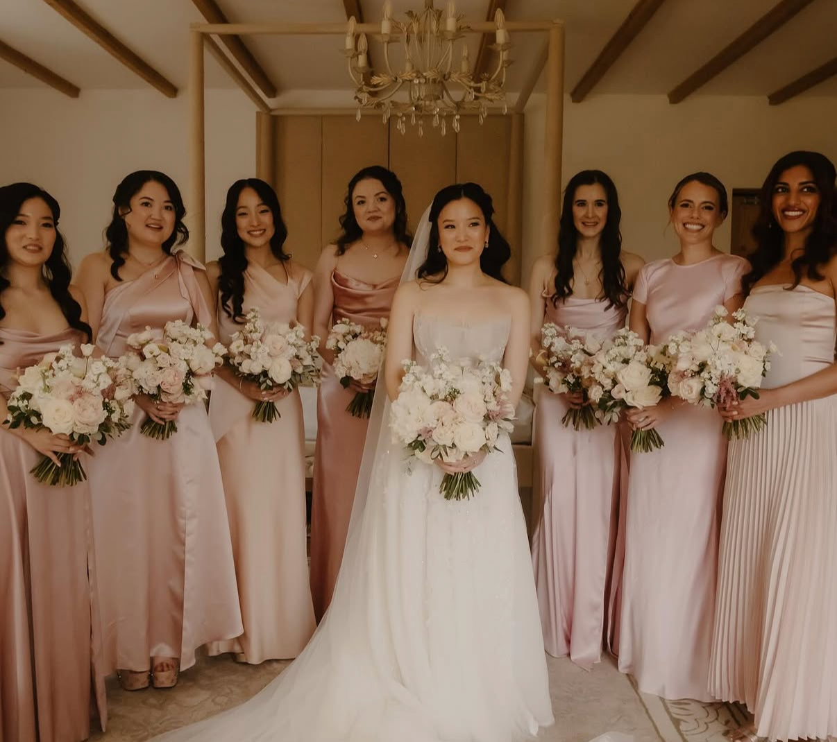 A bride in a wedding dress and eight bridesmaids in pink dresses standing indoors with floral bouquets.