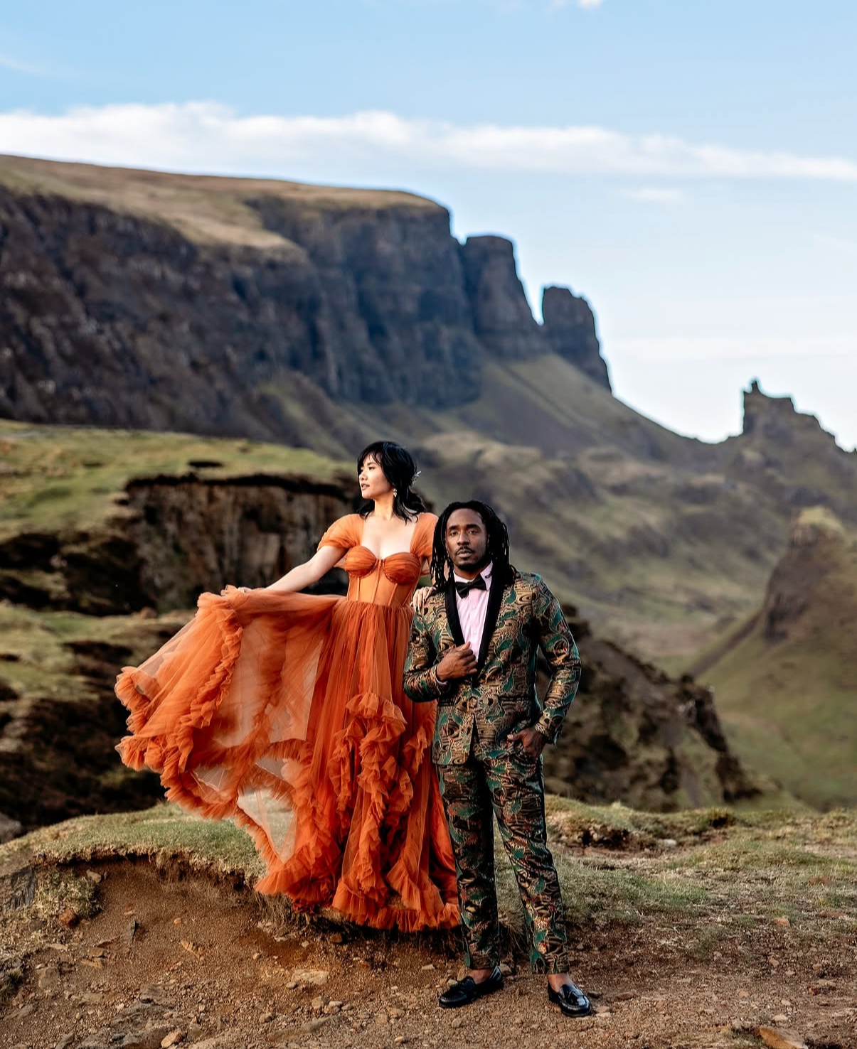 A woman in an orange dress standing on a rock formation, and a man in a patterned suit standing nearby, with a mountainous landscape in the background.