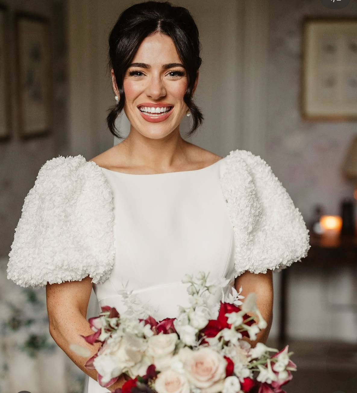 A smiling woman in a white wedding dress holding a bouquet of flowers, standing indoors with blurred background.