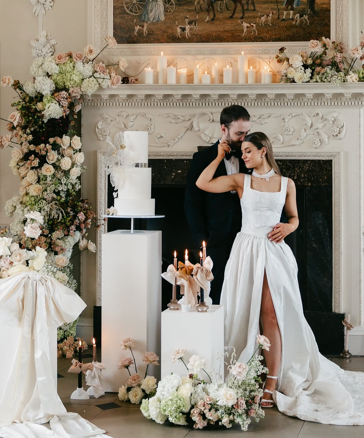 A newlywed couple embraces in front of a decorated fireplace with candles and flowers, celebrating their wedding reception. The bride in a white gown and the groom in a tuxedo stand close together with their eyes closed, surrounded by floral arrangem
