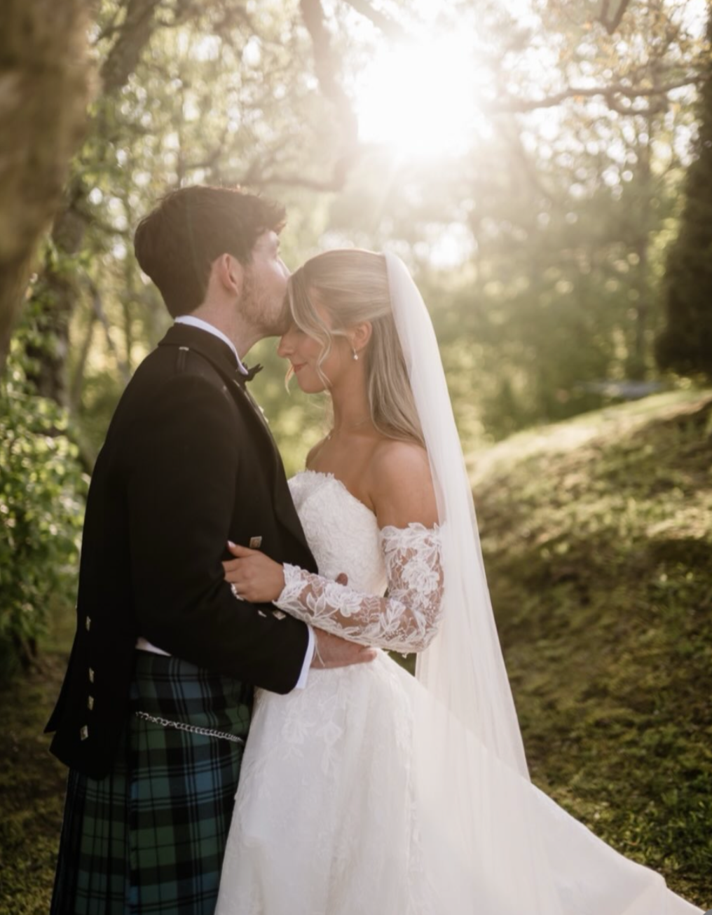 A bride and groom sharing an intimate moment outdoors on their wedding day. The groom is kissing the bride on her forehead, and both are dressed in wedding attire. The background shows sunlight filtering through trees.