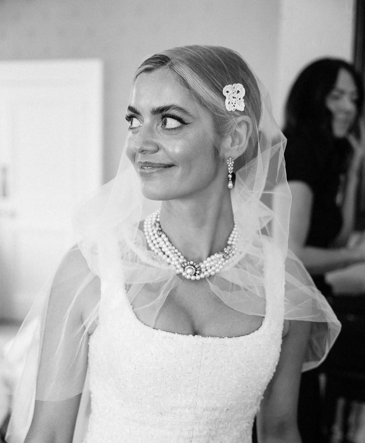 Black and white photo of a woman in a wedding dress, wearing pearls, earrings, a veil, and a flower hair accessory, looking to the side with a smile.