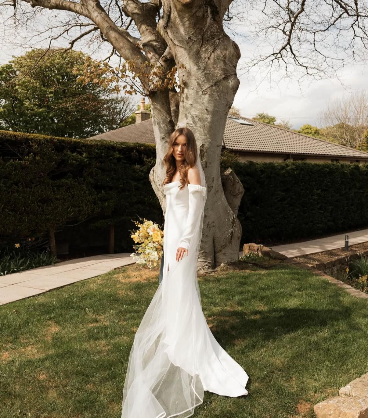 A woman in a white off-the-shoulder wedding dress standing outdoors in front of a large tree, holding a bouquet of flowers, with a house and cloudy sky in the background.