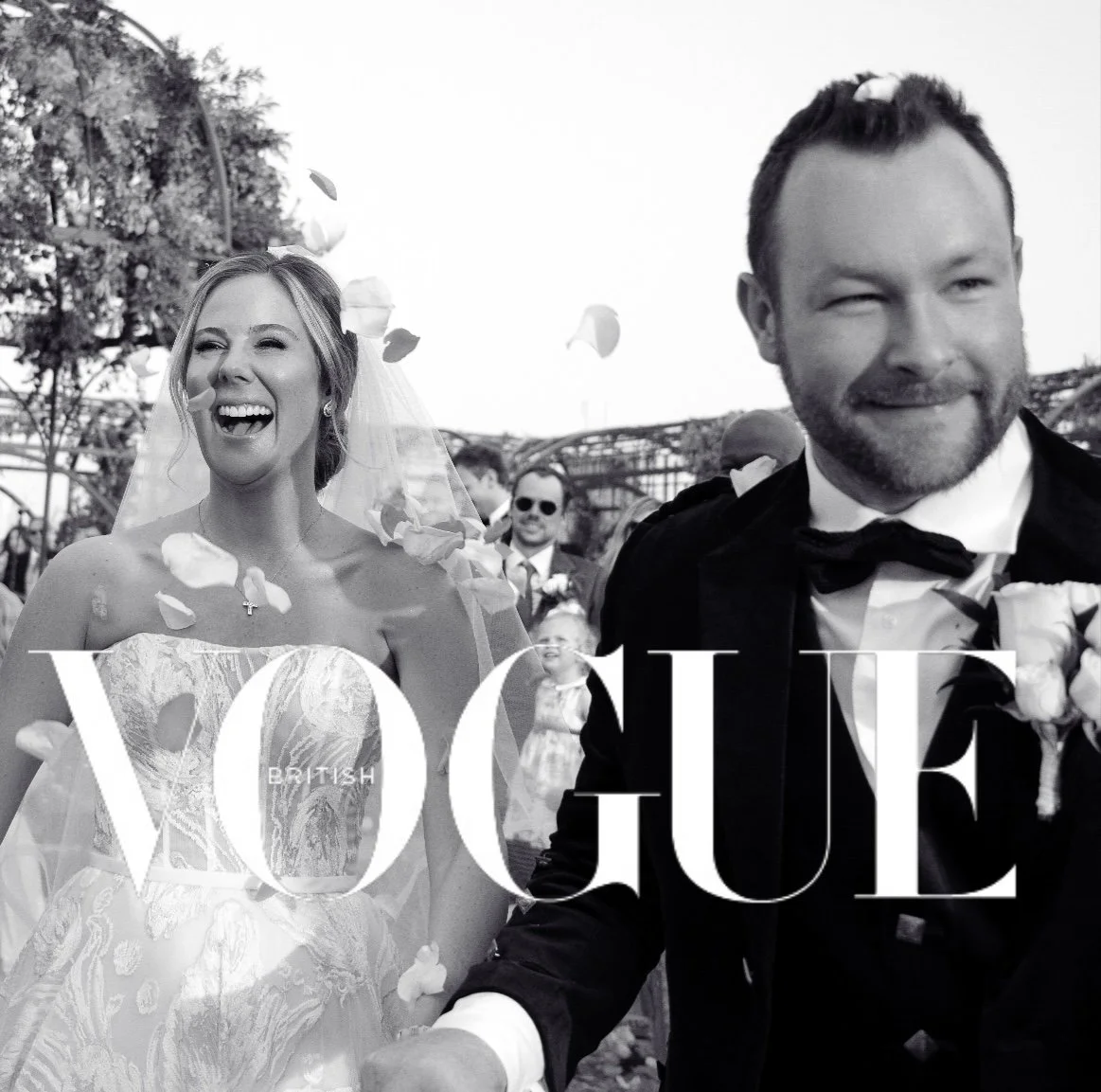 A black-and-white photo of a wedding celebration with a bride in a lace dress and veil, and a groom in a tuxedo, smiling as they walk through falling flower petals, with wedding guests in the background.