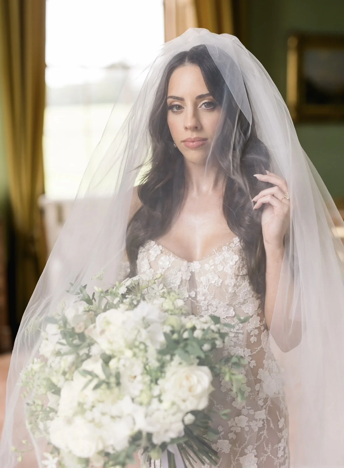 A woman with dark hair wearing a wedding dress and a veil, holding a bouquet of white flowers.