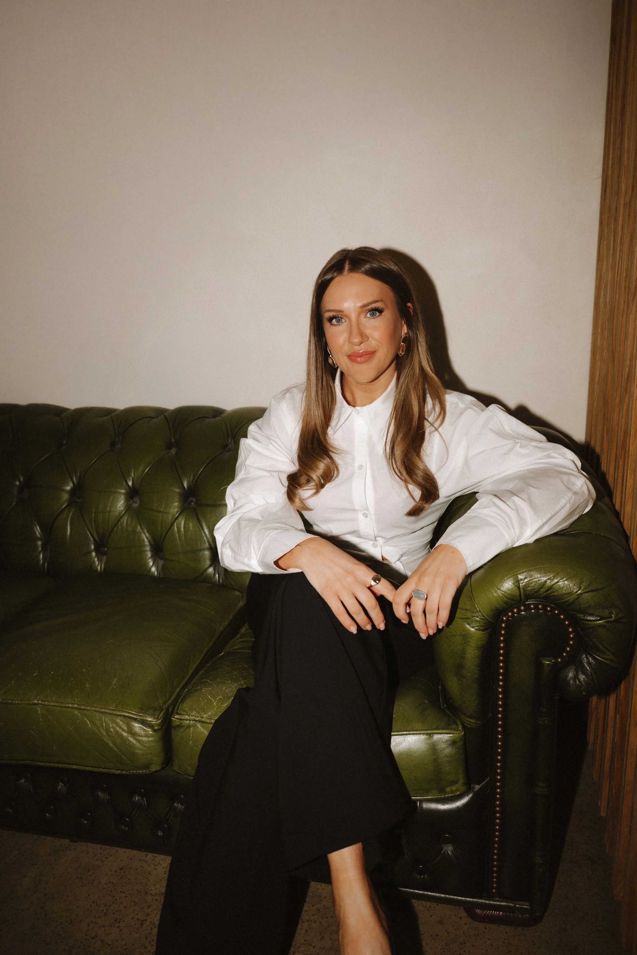 A woman sitting on a green leather tufted sofa, wearing a white blouse and black pants, with long wavy hair and earrings, looking at the camera in a room with a plain wall and wooden panel on the right side.