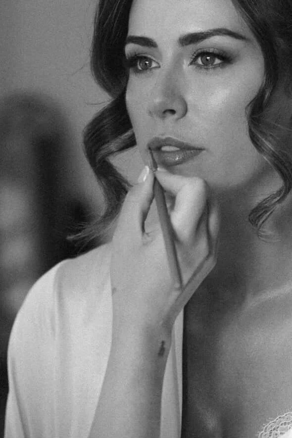 A woman with wavy hair receives makeup applied with a brush on her lips in a black-and-white photograph.