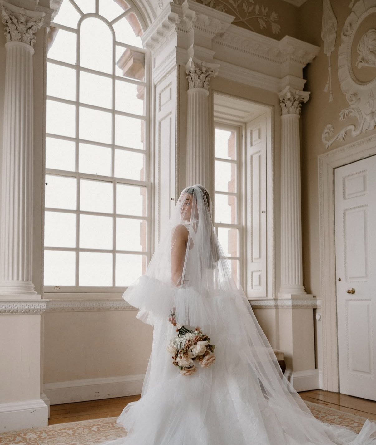 A bride in a white wedding gown and veil standing in front of large windows with ornate columns and decorative trim, holding a bouquet of flowers.