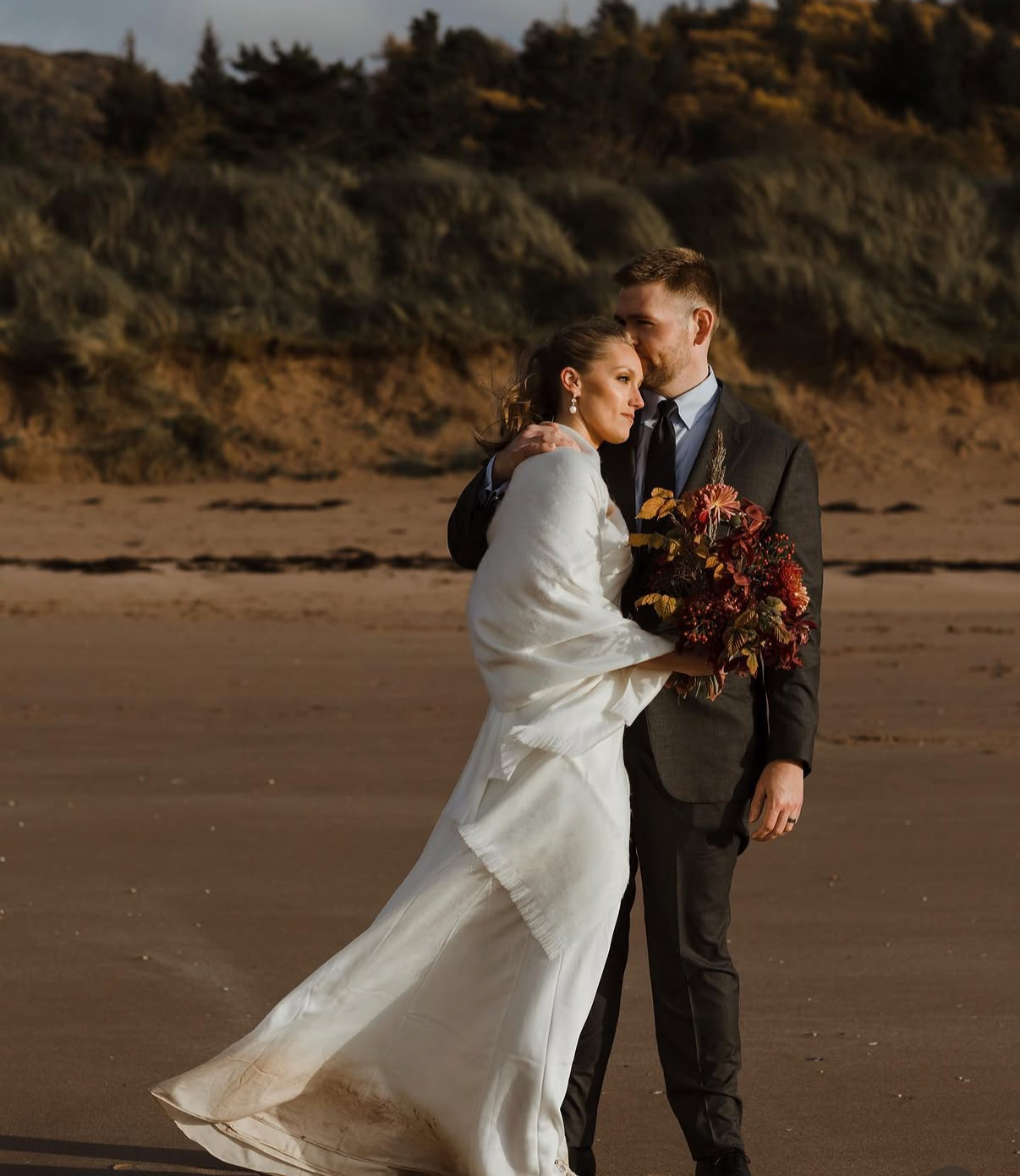 A newlywed couple stands close on a sandy beach with cliffs in the background. The bride wears a white gown and holds a bouquet of red and orange flowers. The groom wears a black suit and gently rests his forehead against the bride's.
