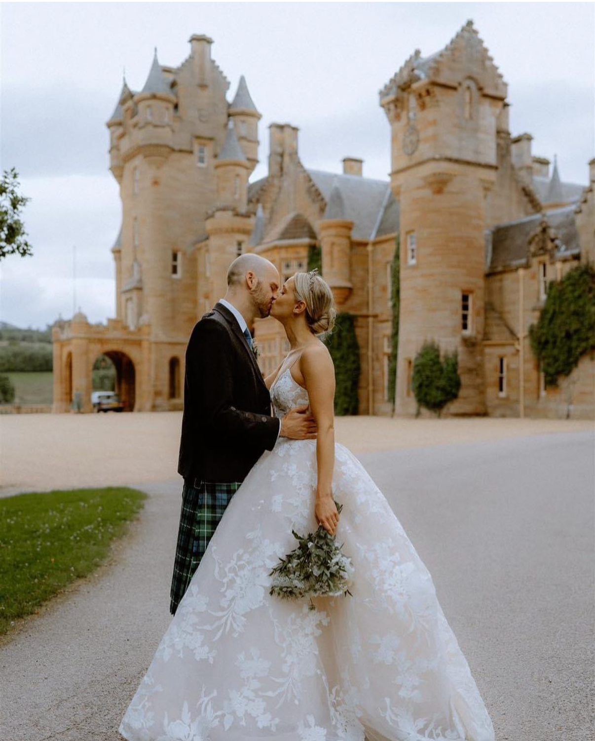 A bride and groom kiss in front of a castle, with the bride holding a bouquet and wearing a lace wedding gown, and the groom in a tartan kilt and jacket.
