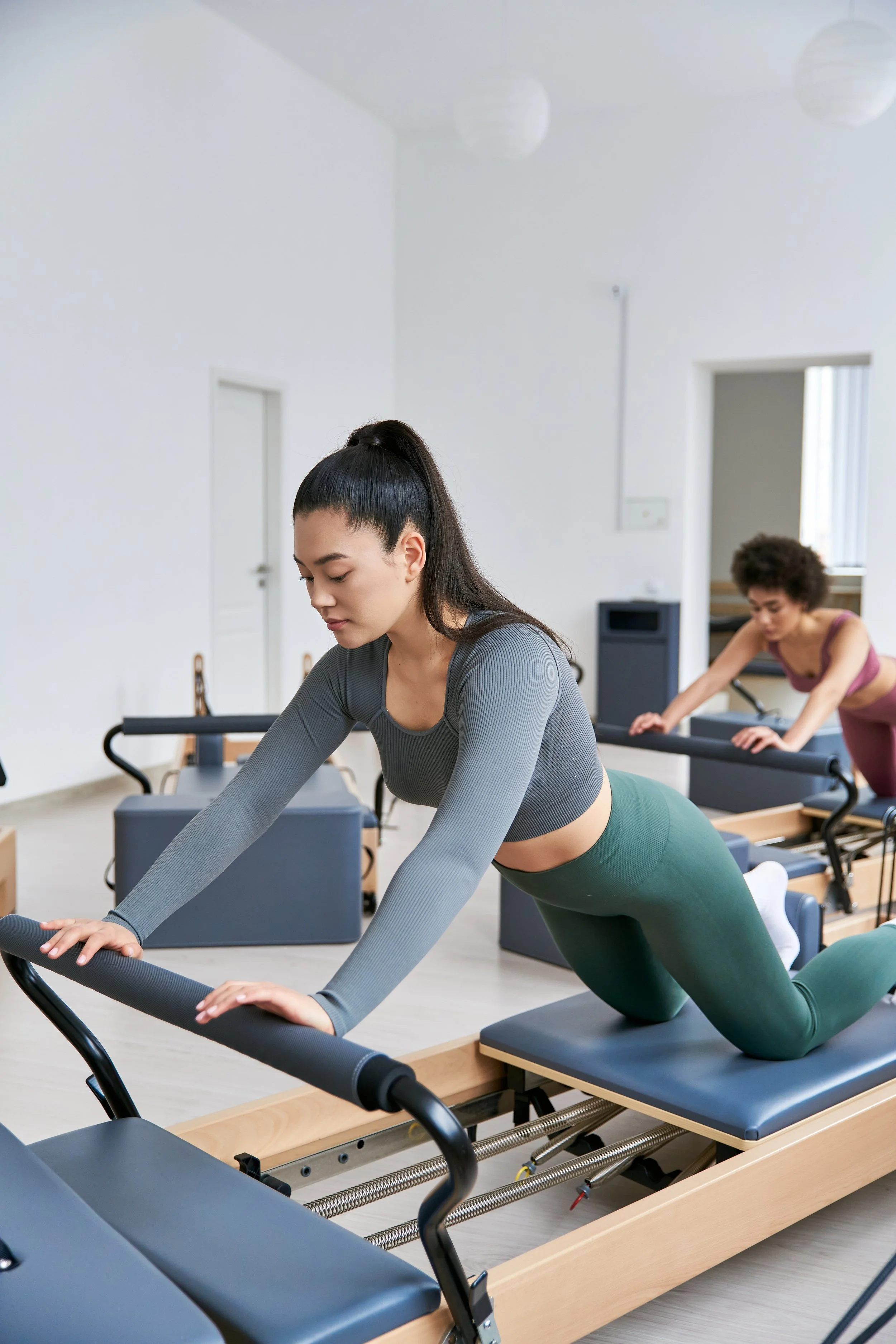 Two women are participating in a Pilates class, using reformer exercise machines in a bright, white-walled studio. The woman in the foreground has dark hair in a high ponytail, is wearing a gray long-sleeve top and green leggings, and is in a kneeling position on the reformer with her hands on the parallel bars. The woman in the background has curly hair, wears a pinkish workout top, and is also engaged in an exercise on a reformer machine.