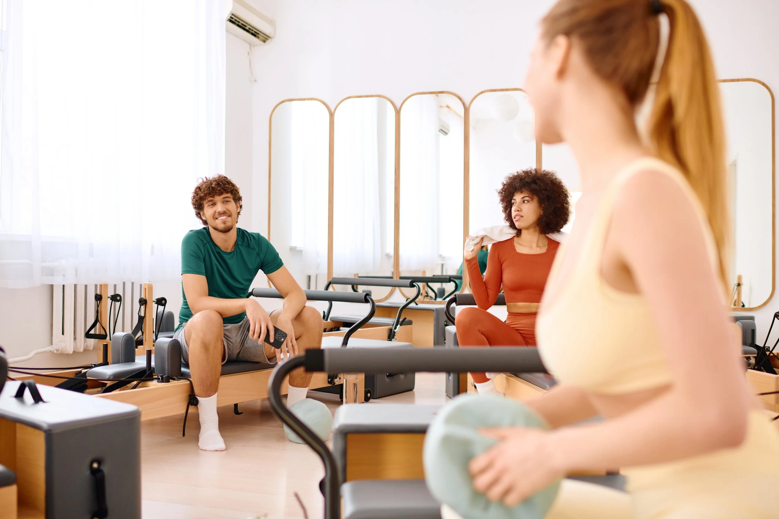 Two women and a man in a fitness class using Pilates reformer machines in a bright studio with large windows and mirrors.