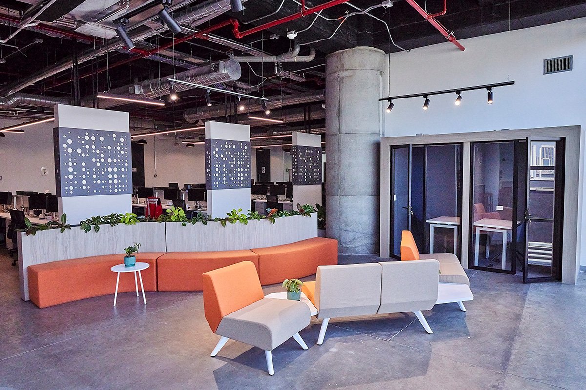 Área de descanso moderna con sillones naranjas y beige, mesas con plantas y separadores con diseño de círculos blancos, en un espacio con estructura de cemento y ductos expuestos en el techo.