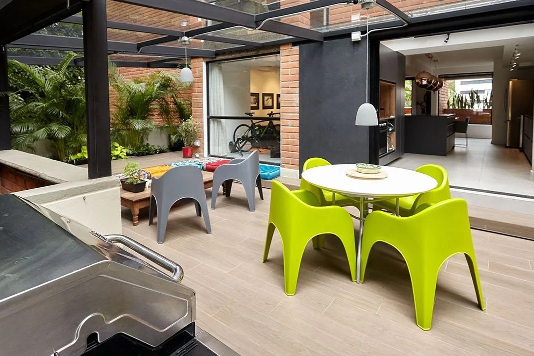 Zona de comedor con mesa redonda y sillas verdes en un espacio interior con vista a un patio con plantas y muebles para descansar.
