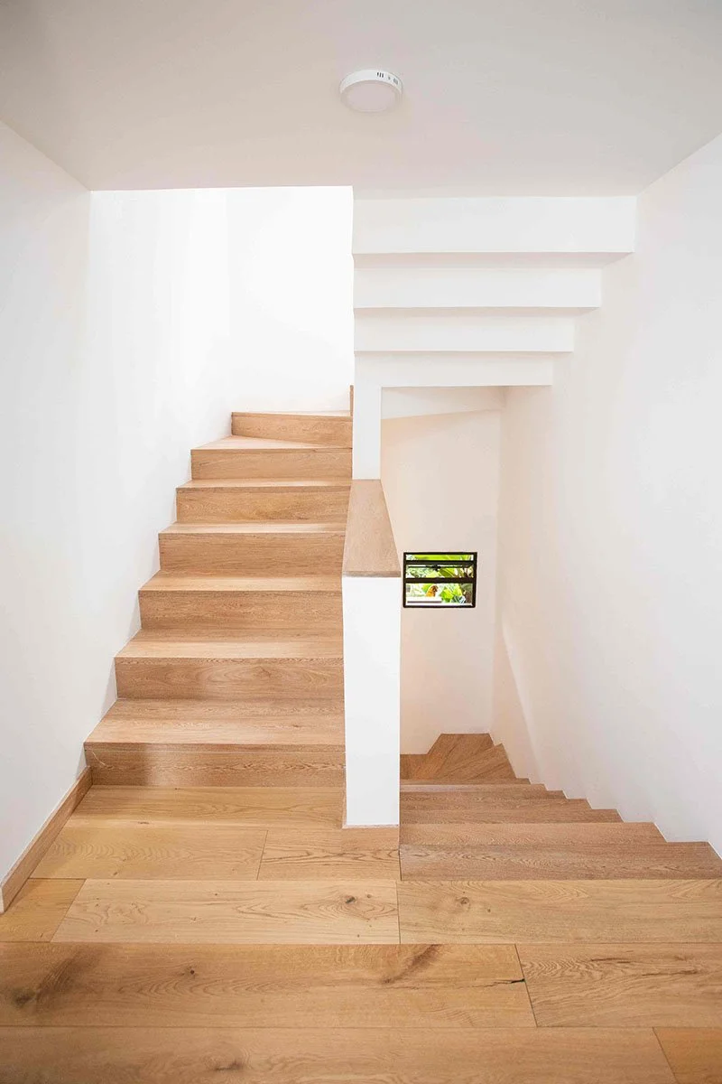 Escaleras de madera en una casa moderna con paredes blancas y una pequeña ventana con vista al exterior.