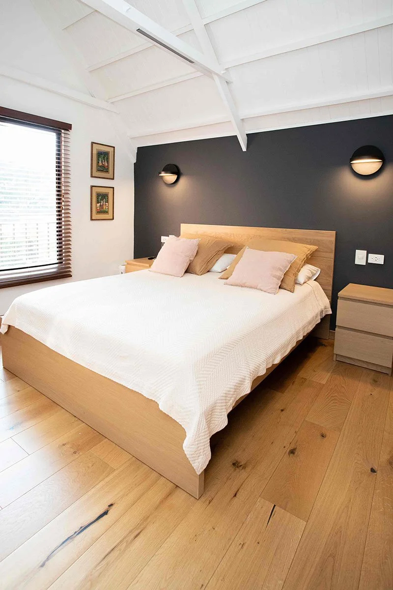 Cama grande con sábanas blancas y almohadas rosadas y beige, con dos mesitas de noche de madera a ambos lados, en una habitación con pared negra, ventanas con contraventanas de madera y cuadros en la pared, y techo blanco con vigas.