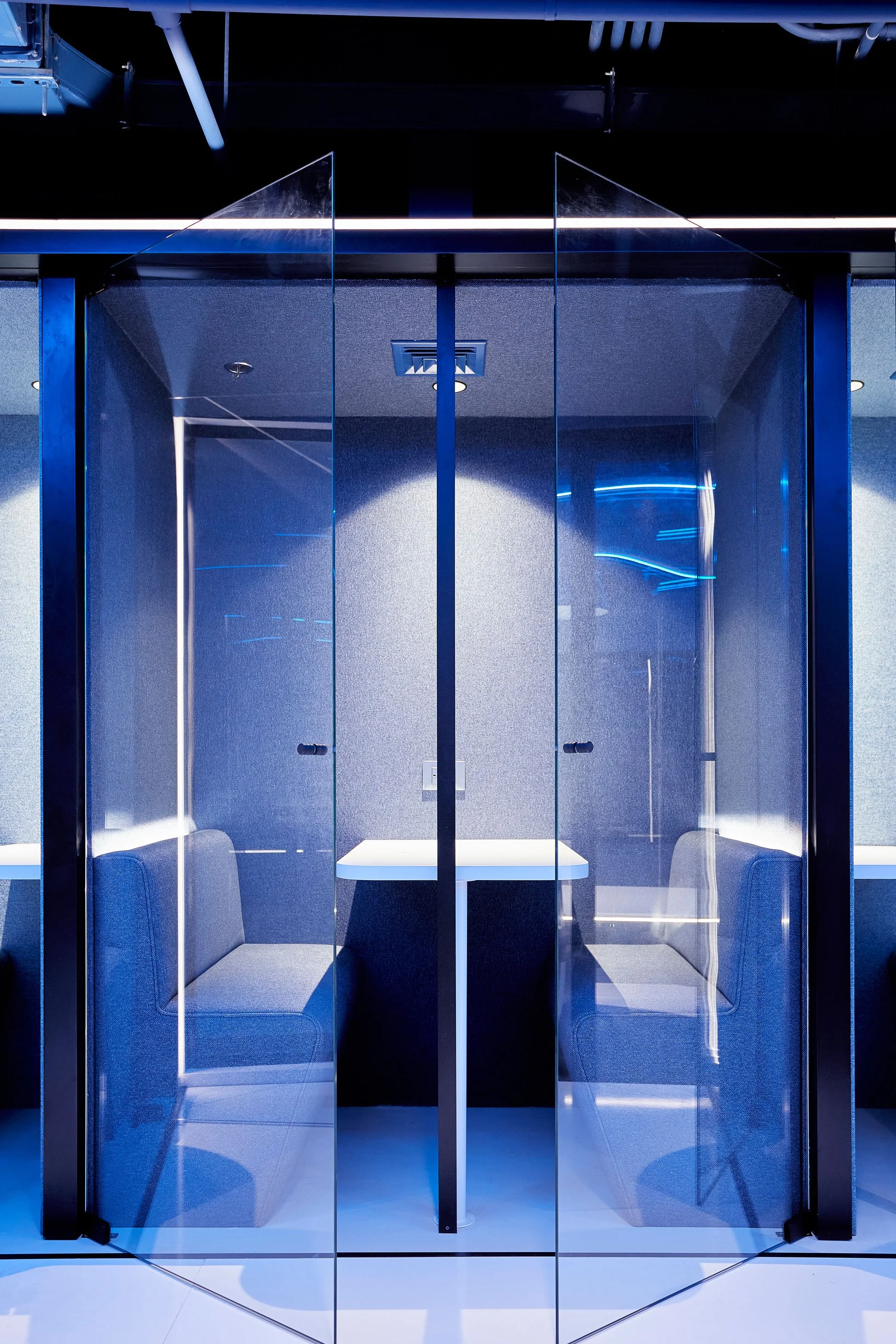 Una sala de reuniones con paredes de vidrio, hay una mesa en el centro, sillas a los lados y un ambiente moderno con iluminación azul.