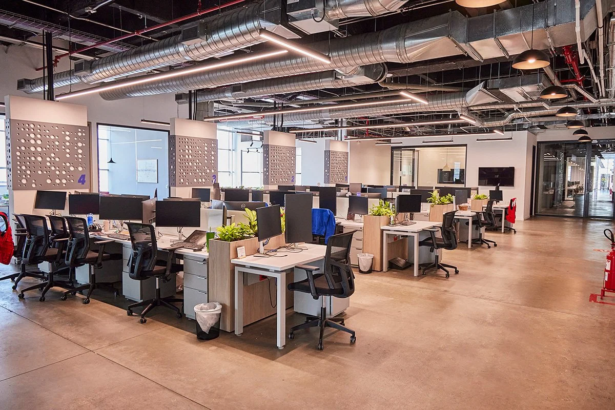 Oficina moderna con escritorios, sillas, monitores y plantas, en un espacio abierto con iluminación natural y ventilación industrial en el techo.