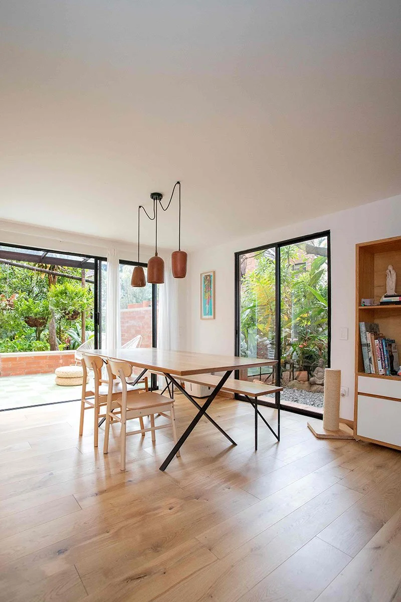 Sala comedor luminosa con mesa de madera y sillas, ventanas grandes con vista a un jardín, lámparas colgantes de color terracota, estantería con libros y objetos decorativos.