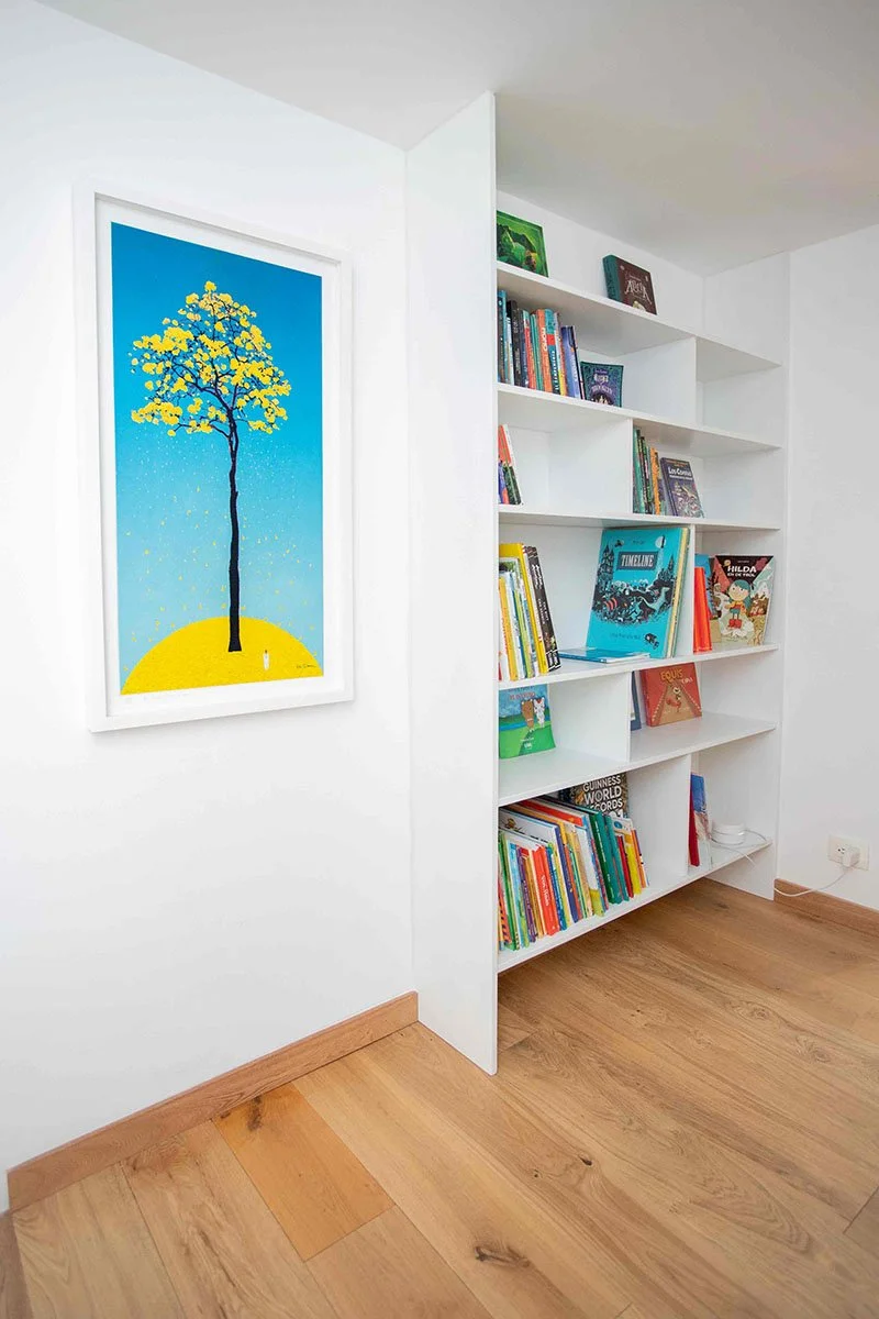 Estantería blanca llena de libros en una pared blanca junto a un cuadro con un árbol y un cielo azul.