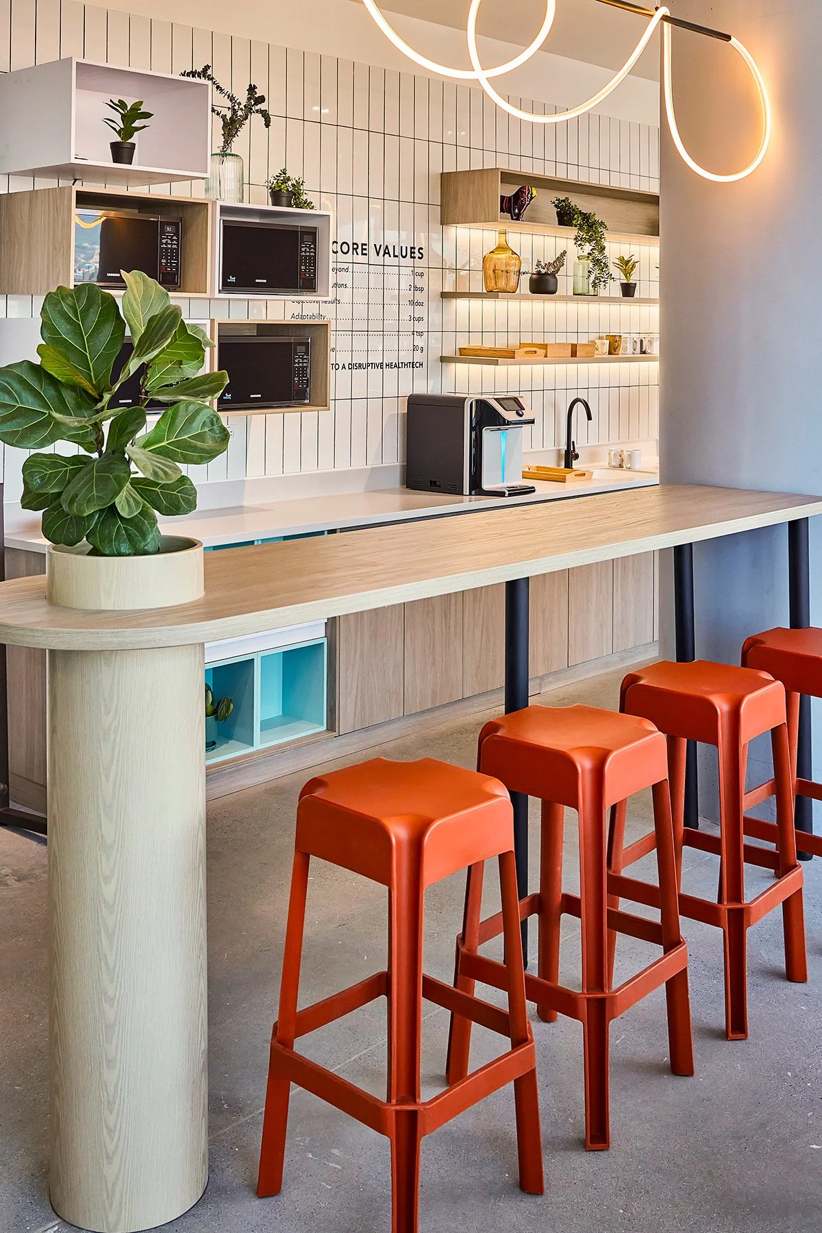 Una barra de cocina moderna y minimalista con pufs naranja, mesa de madera clara, plantas en macetas, microondas y una cafetera. Paredes blancas con azulejos y estanterías con utensilios y decoraciones.