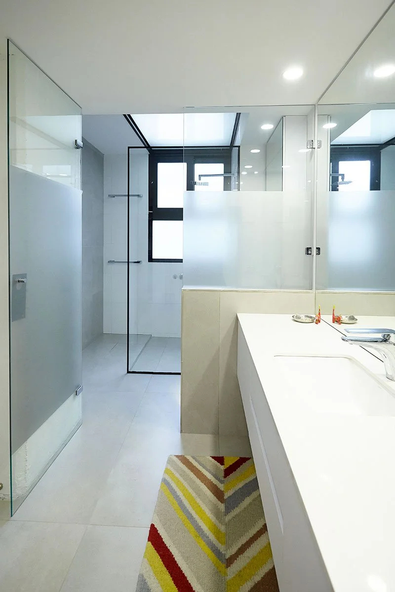 Baño moderno con ducha separada por pared de vidrio, lavabo con espejo grande, ventanas, iluminación empotrada y alfombra con rayas de colores.