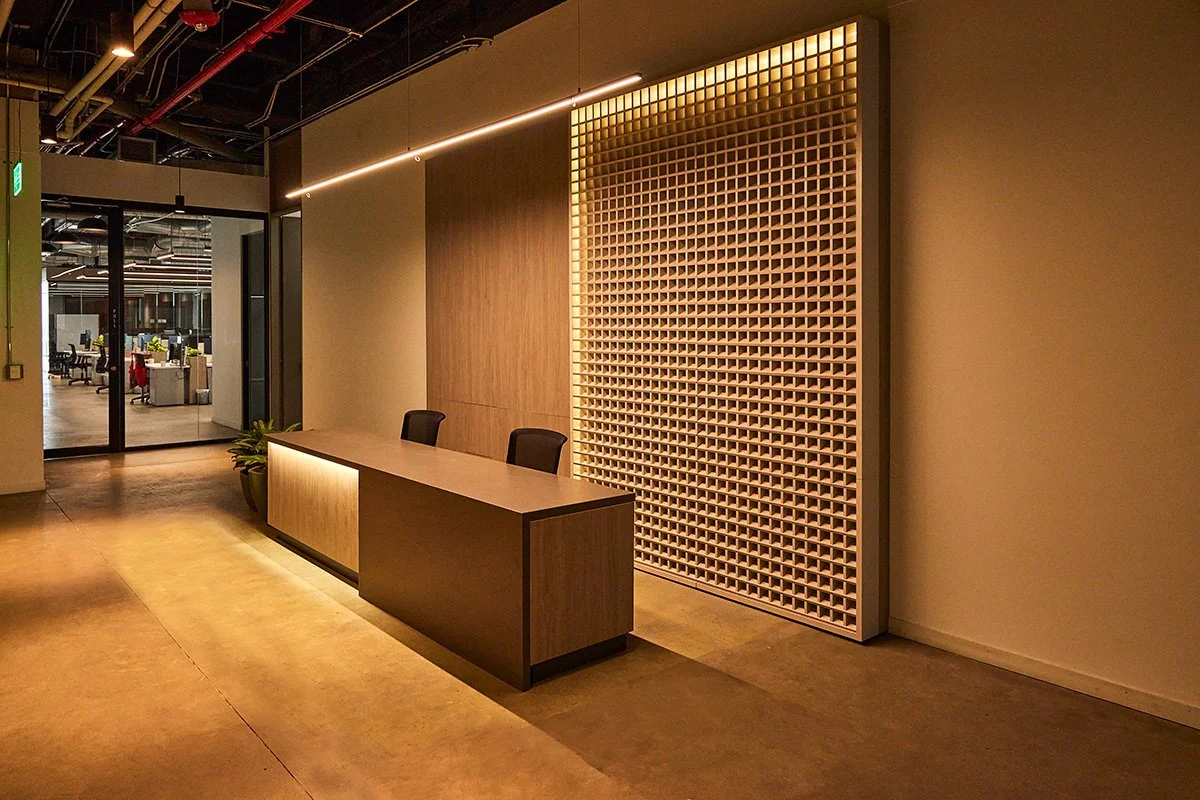 Mostrador de recepción con dos sillas negras frente a una pared con decoración de panel de madera y iluminación en un espacio interior de oficina moderna