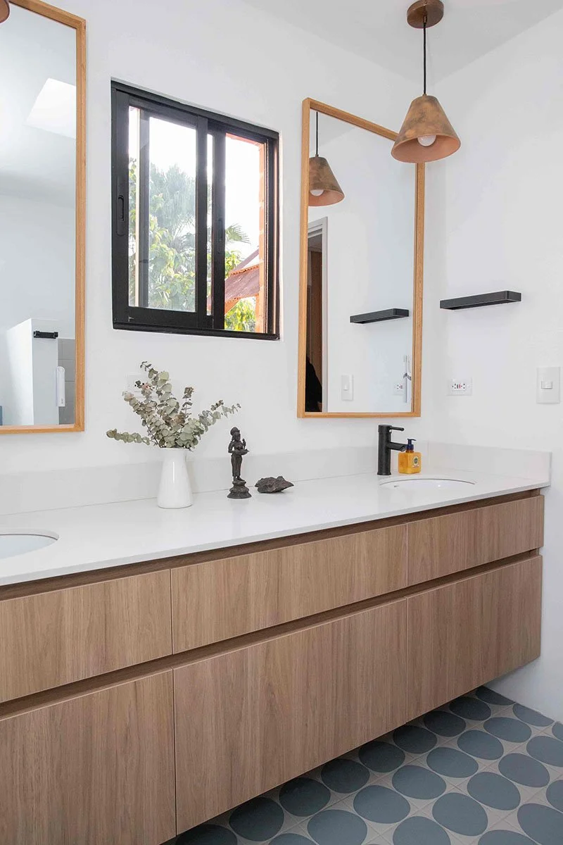 Lavabo de tocador de madera con espejo grande y ventana a la izquierda. Decoraciones de esculturas y plantas, con lámparas colgantes de tono marrón.