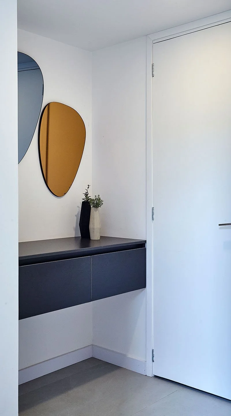 Entrada a una habitación con pared blanca, espejo decorativo de formas orgánicas en tonos gris y mate, un mueble oscuro con dos cajones y una planta en un jarrón en la esquina de la superficie.