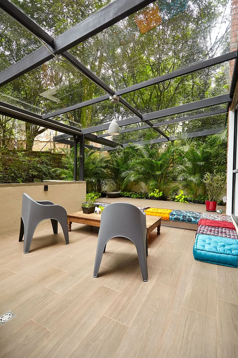 Un espacio de terraza con techo de vidrio, plantas verdes y muebles de madera y cojines coloridos.