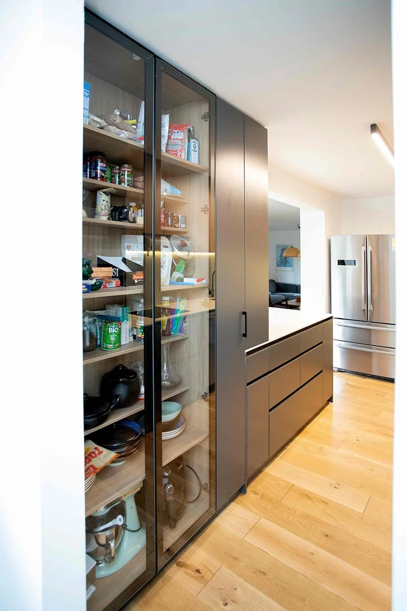 Gabinete de cocina con puertas de vidrio que muestra alimentos y utensilios, en una cocina moderna con electrodomésticos de acero inoxidable y suelo de madera.